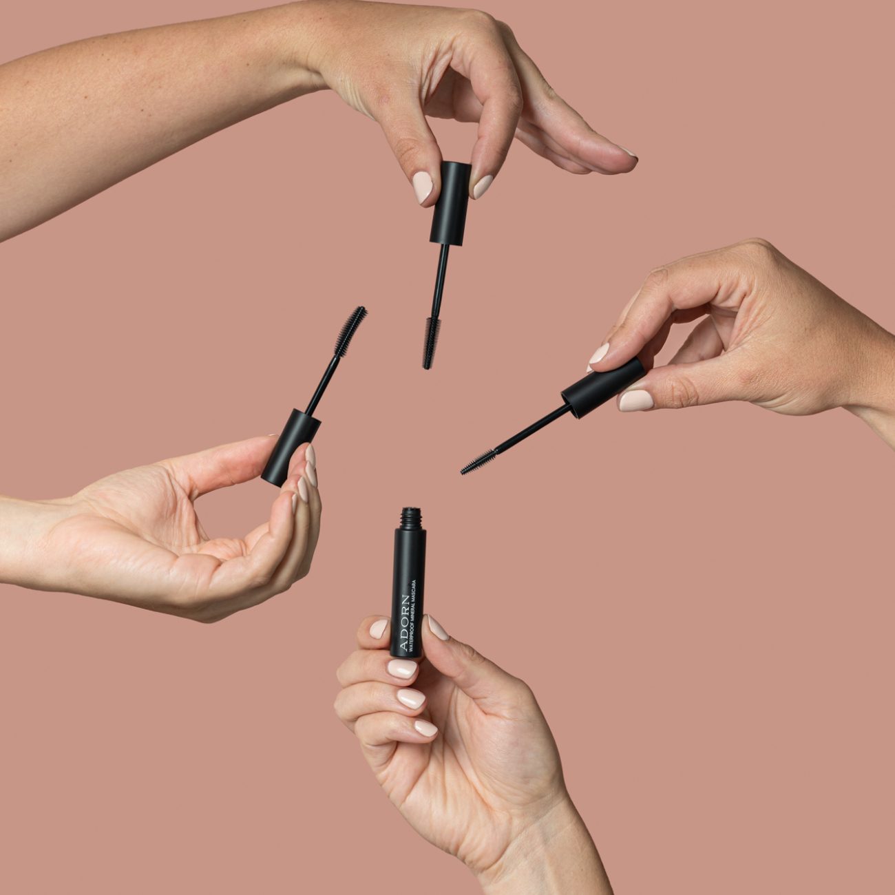 Hands holding mascara wands around an Adorn Cosmetics mascara tube in a styled beauty product photography setup.
