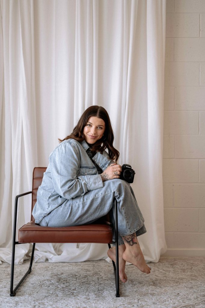 Woman sitting on a chair holding a camera during a natural light personal brand photography session in a studio setting.