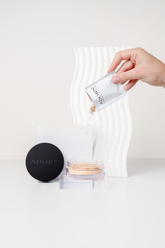 Hand pouring loose mineral makeup into an open Adorn Cosmetics jar in a minimalist studio product photography setup.