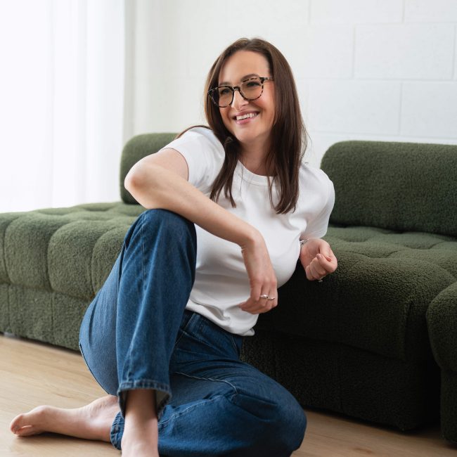 Studio portrait of a woman wearing a white top and jeans, seated on the floor and leaning against a green sofa, smiling in a bright studio setting.