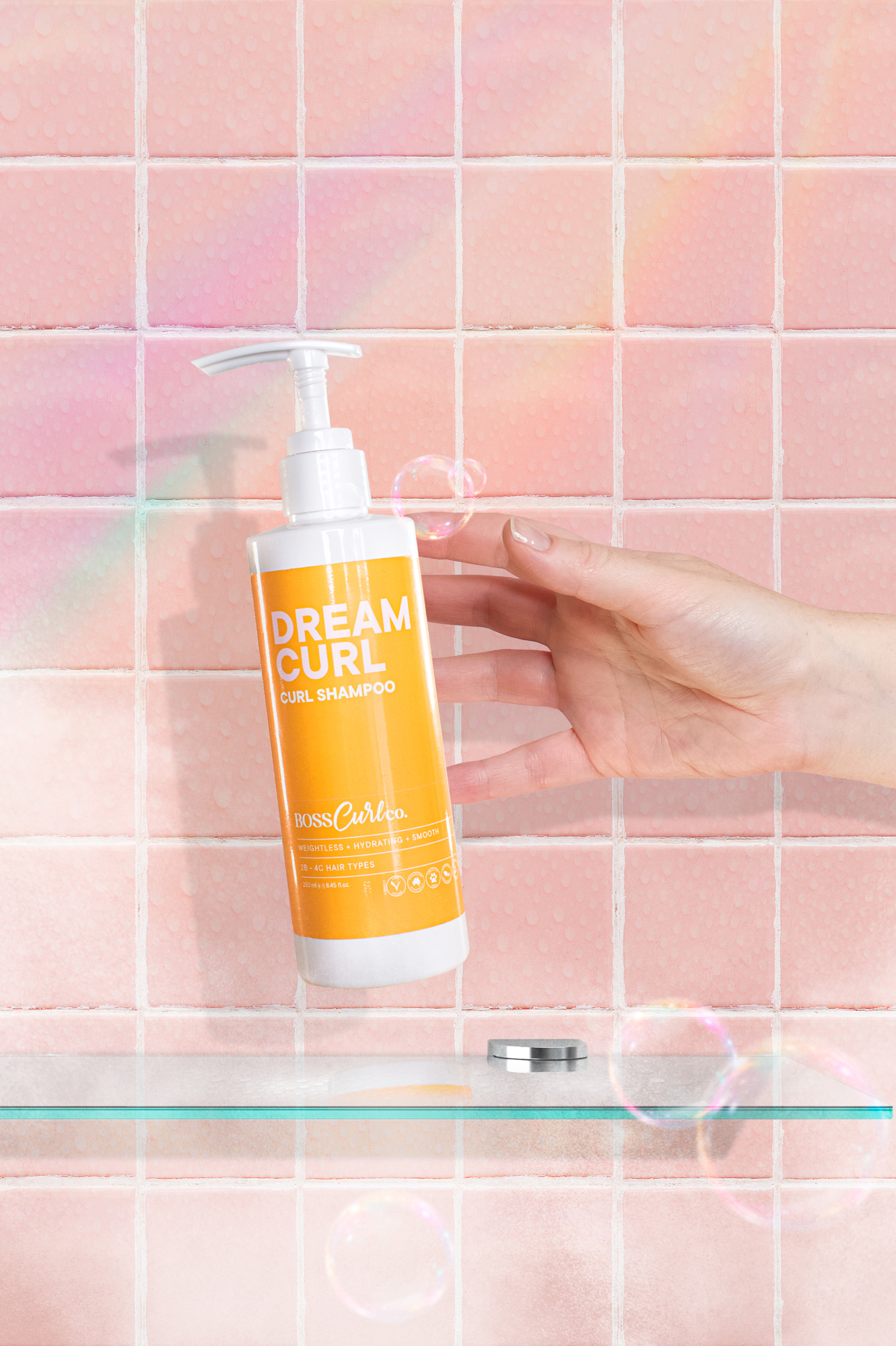 Dream Curl shampoo bottle held in hand against pink tiled bathroom background with bubbles