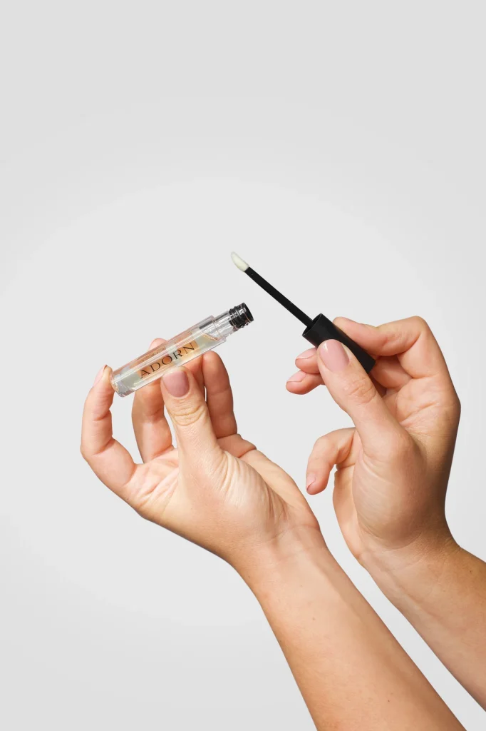 Two hands holding an Adorn lip gloss tube and applicator wand against a white background