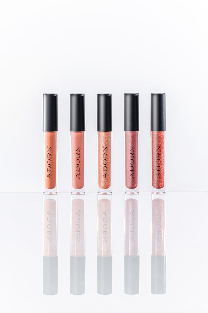 Five Adorn lip gloss shades standing in a row on a white reflective surface