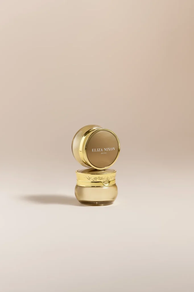 Two stacked gold Eliza Nixon Brows pots on a warm neutral background