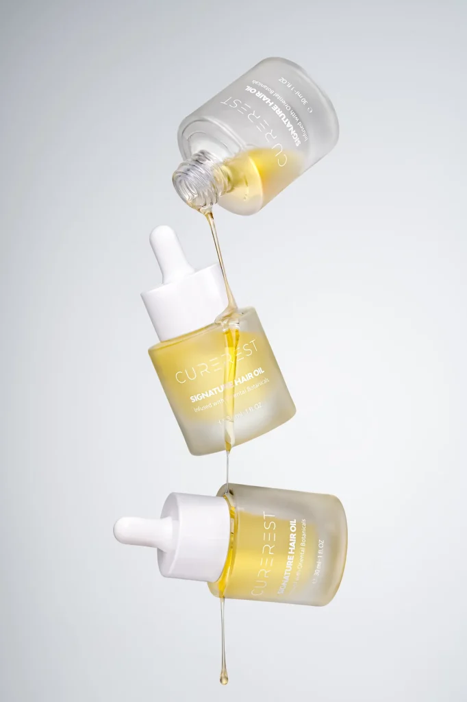 Three Curerest Signature Hair Oil bottles suspended with oil pouring between them on a soft grey background