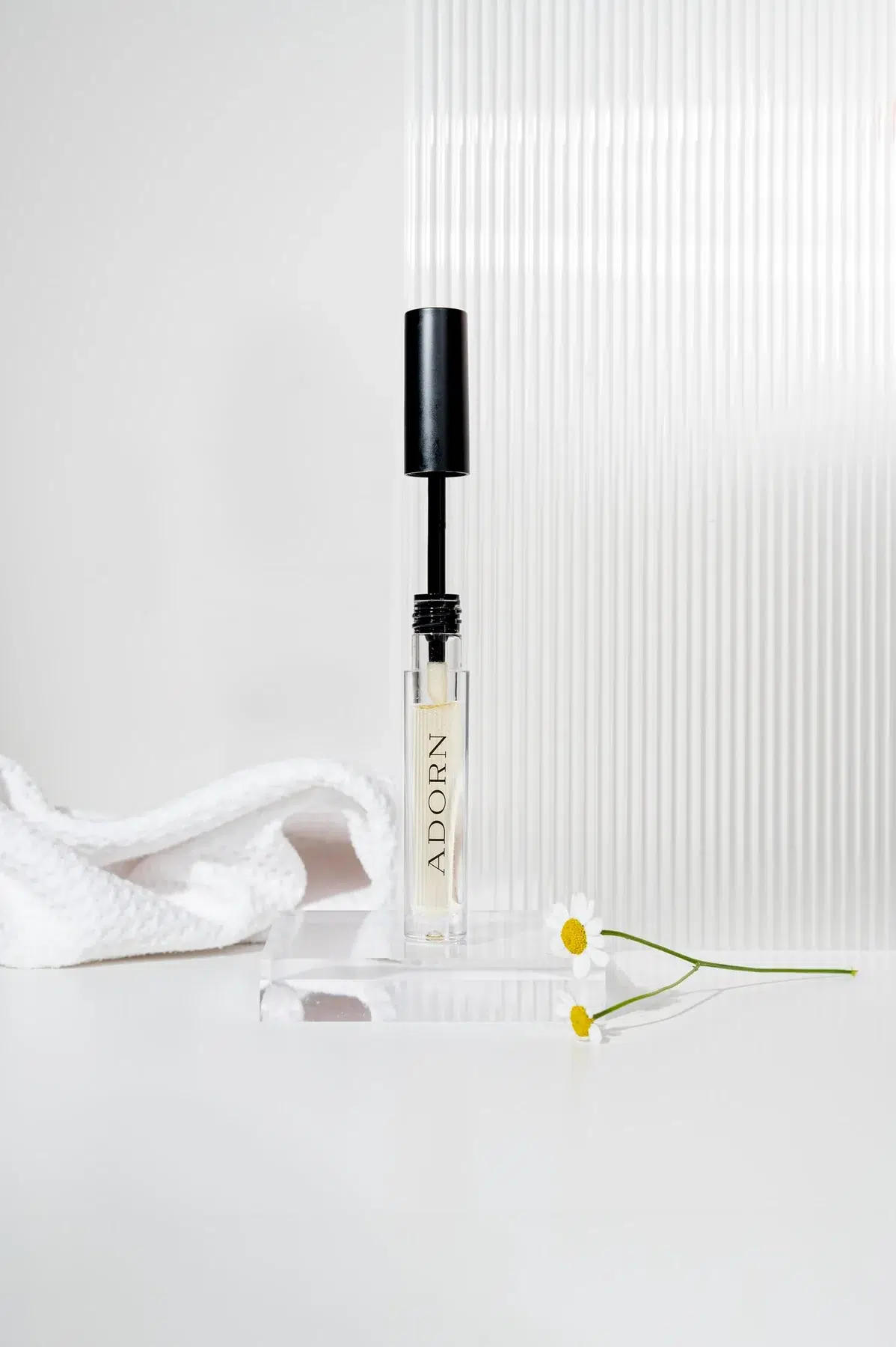 Adorn serum vial with wand applicator on an acrylic block with daisies and a white towel