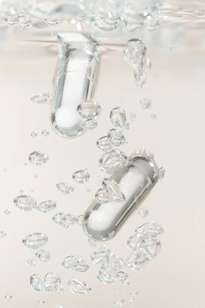 Two clear Genetic Labs supplement capsules sinking through water with rising bubbles