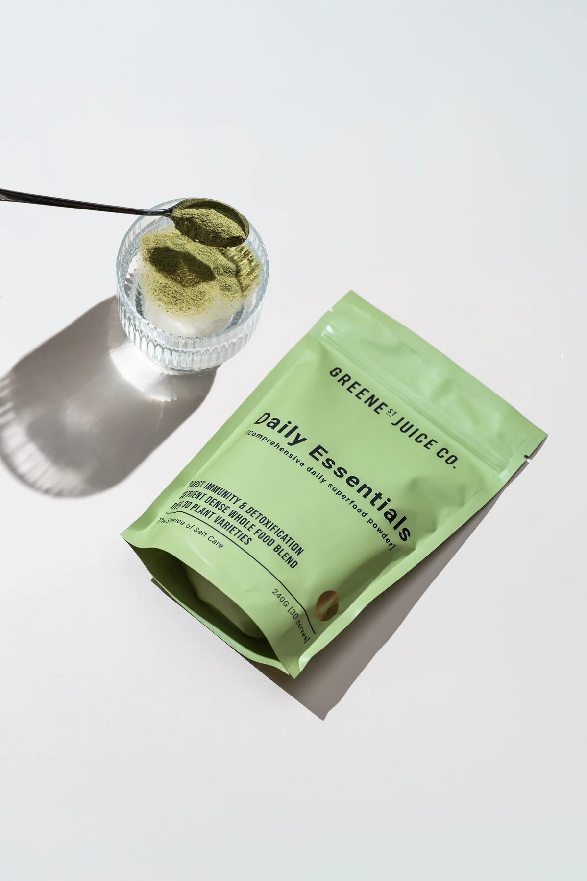 Greene Street Juice Co Daily Essentials pouch beside a glass of green powder and a spoon on white