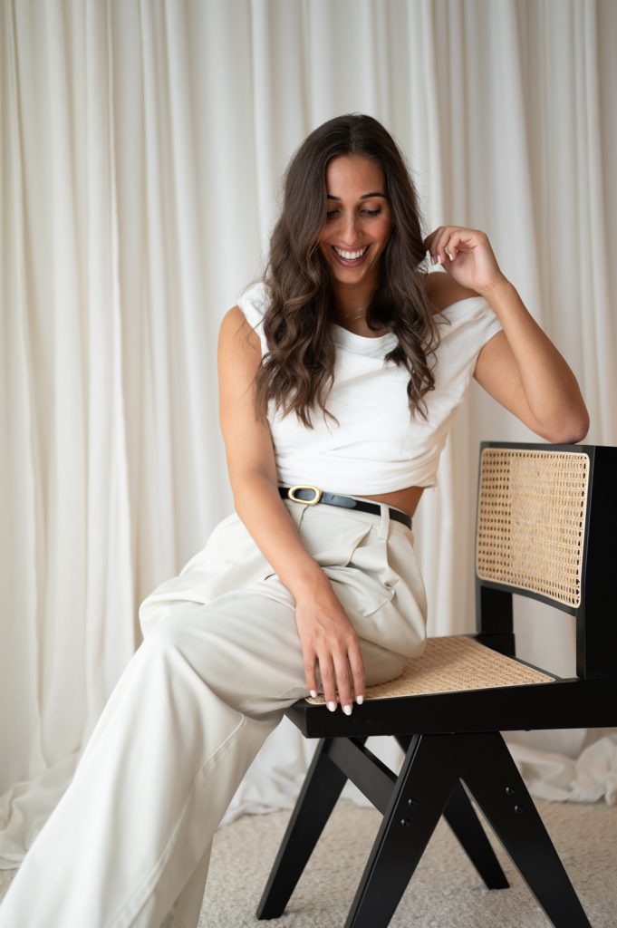 Business owner Emily sitting on a black chair in a light-filled photography studio, smiling while looking down.