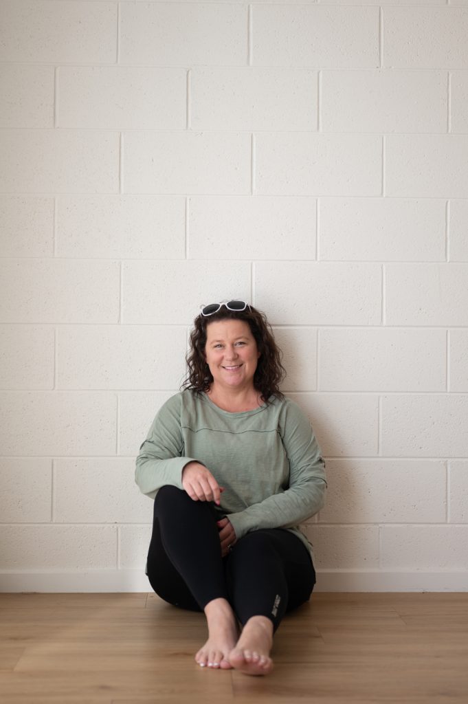 Business owner Leanne sitting barefoot against a white brick wall, smiling at the camera during a portrait session.
